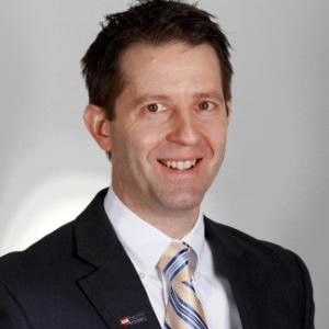 Professional headshot of a man in a dark suit and striped tie, smiling at the camera.