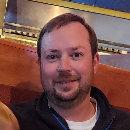 Portrait of a smiling man with short brown hair and a beard, wearing a dark jacket, sitting in a booth indoors.