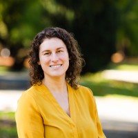 Portrait of a smiling woman with curly brown hair wearing a yellow blouse, outdoors in a sunny park.