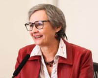 Smiling woman with short gray hair and glasses speaks into a microphone, wearing a red leather jacket and white collared shirt.