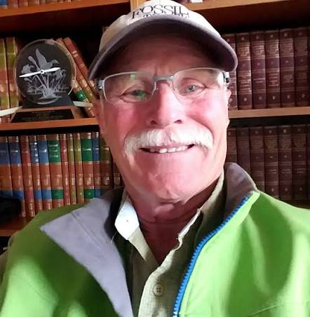 Smiling older man wearing a cap and glasses in a library with books on shelves behind him.