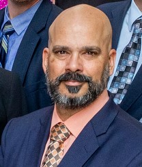 Close-up of a bald man with a beard wearing a navy suit, peach shirt, and patterned tie in a formal group photo.