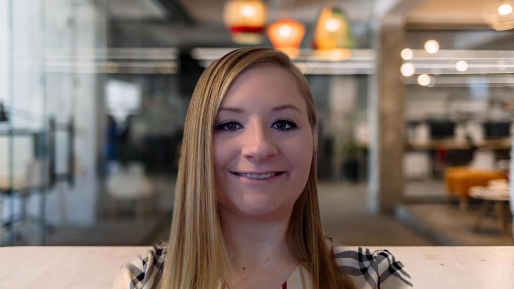 Smiling woman with long blond hair sits at a light desk in a modern open-plan office.