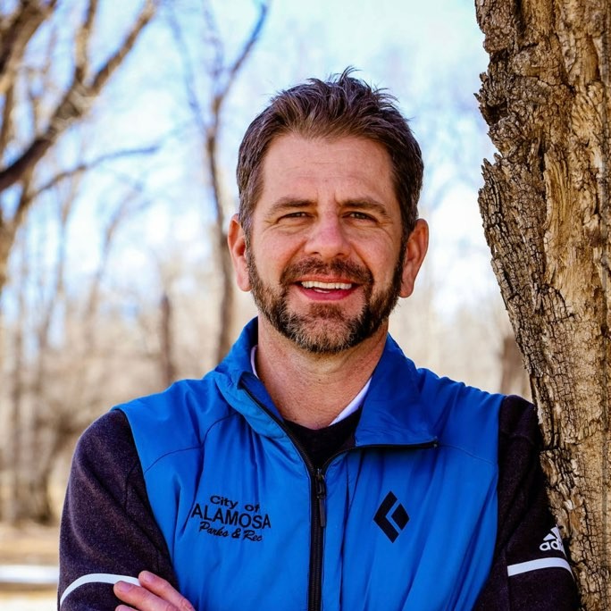 Man smiling outdoors, wearing a blue vest with City of Alamosa Parks & Rec logo, leaning against a tree.