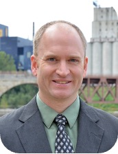 Smiling man in a gray suit and green shirt with a patterned tie, outdoors near a river and city buildings.