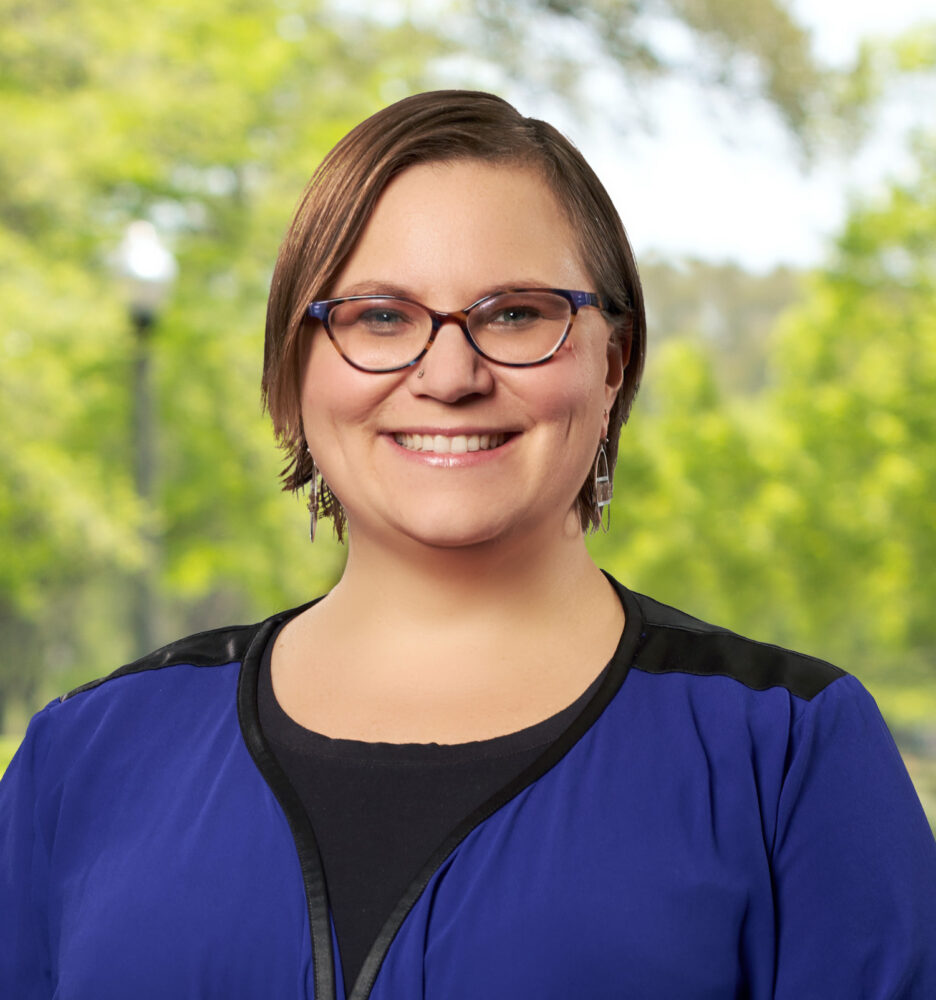 Smiling woman with short brown hair and glasses outdoors, wearing a blue top, blurred green background