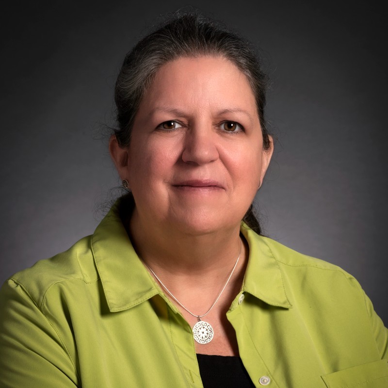 Woman with gray hair in a lime green shirt, facing the camera against a gray backdrop, wearing a silver pendant necklace.
