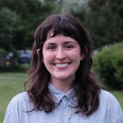 Young woman with brown hair in a light blue striped button-down shirt, smiling outdoors with greenery behind.