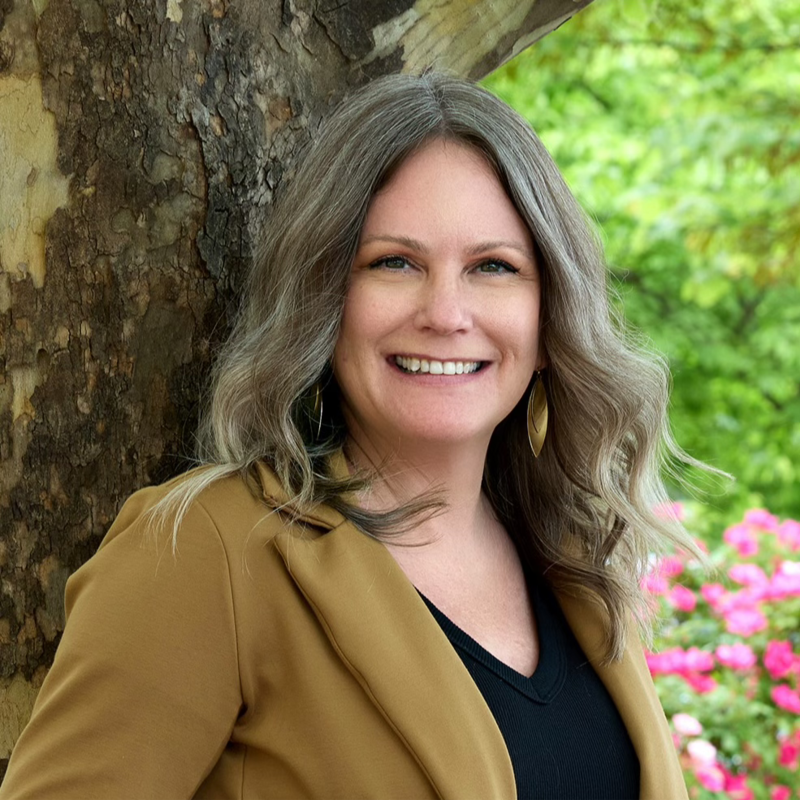 Smiling woman with wavy gray-blonde hair, wearing a tan blazer and black top, leaning against a tree in a garden.