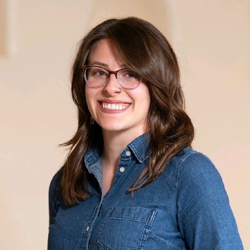 Smiling woman with shoulder-length brown hair and glasses in a denim shirt against a neutral background, facing slightly to the left.