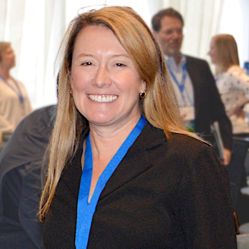 Smiling woman in a black blazer with a blue lanyard at a conference or networking event, facing the camera.