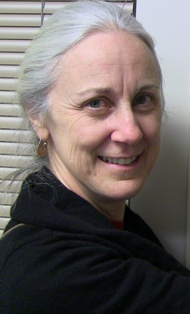 Smiling older woman with gray hair tied back, wearing a black sweater, leaning against a wall with horizontal blinds behind her.