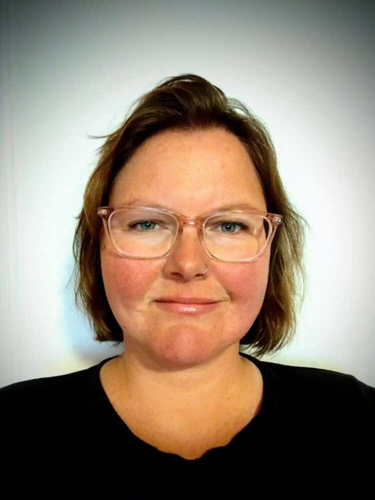 Close-up portrait of a woman with short brown hair and pink-framed glasses, looking at the camera against a pale background.