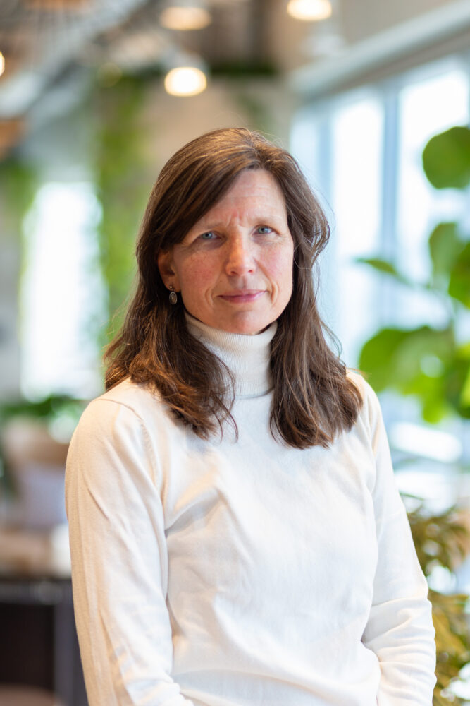 Portrait of a woman with shoulder-length brown hair wearing a white turtleneck, standing in a bright indoor space with plants in the background, looking at the camera.