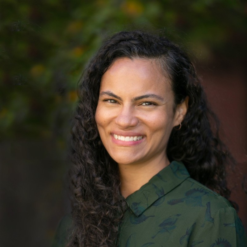 Portrait of a smiling woman with curly dark hair outdoors, wearing a green patterned shirt.