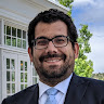 Professional portrait of a smiling man with dark hair, beard, and glasses in a blue suit, outdoors near a white building.