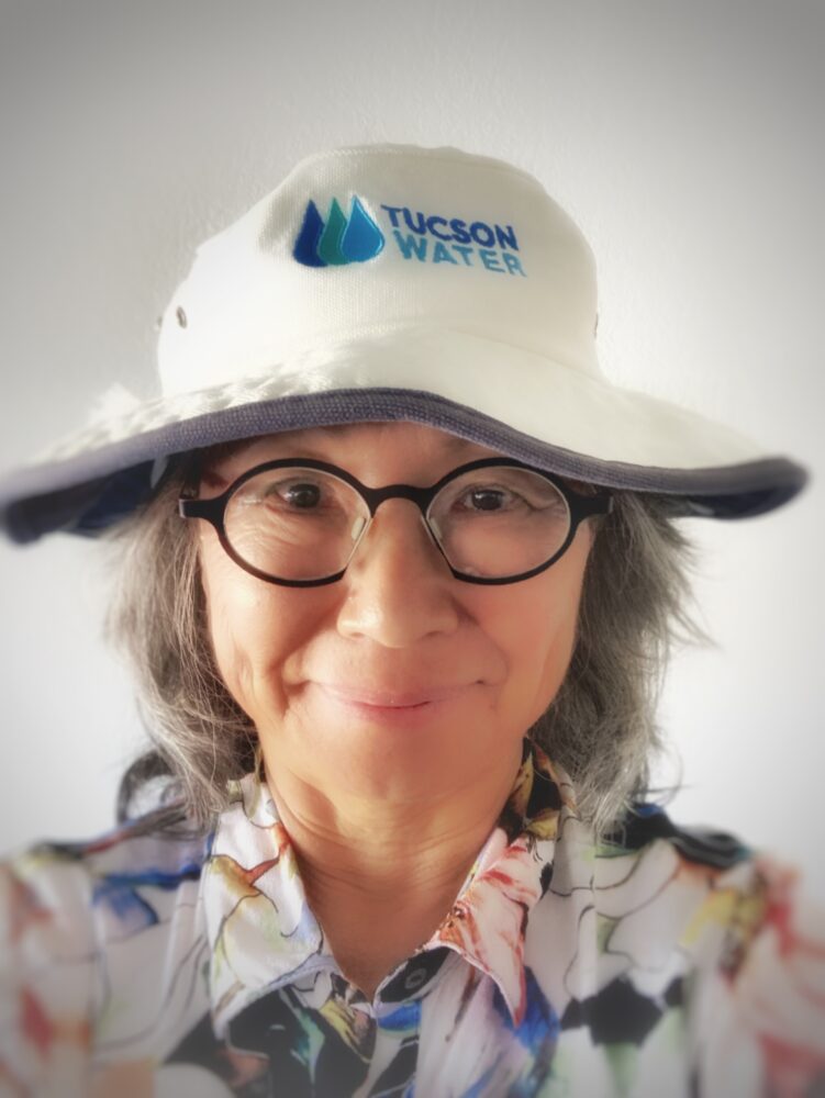 Woman wearing a white Tucson Water hat with blue-green logo, glasses, and a colorful floral shirt, smiling at the camera