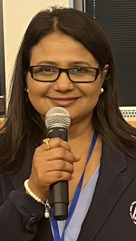 Woman with glasses and pearl earrings speaks into a handheld microphone at a formal event, wearing a navy blazer and light blue blouse with a blue lanyard.
