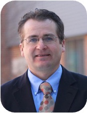 Portrait of a middle-aged man wearing glasses, a navy suit and an orange patterned tie, standing outdoors with a brick building behind.