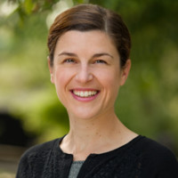 Smiling woman with short brown hair wearing a black sweater, outdoors with greenery in the background.