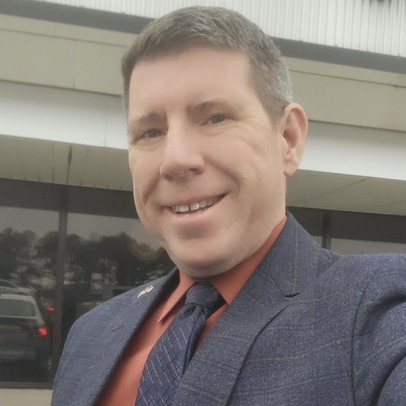 Head-and-shoulders shot of a man in a blue checked blazer and red shirt smiling outdoors, taking a selfie outside a building