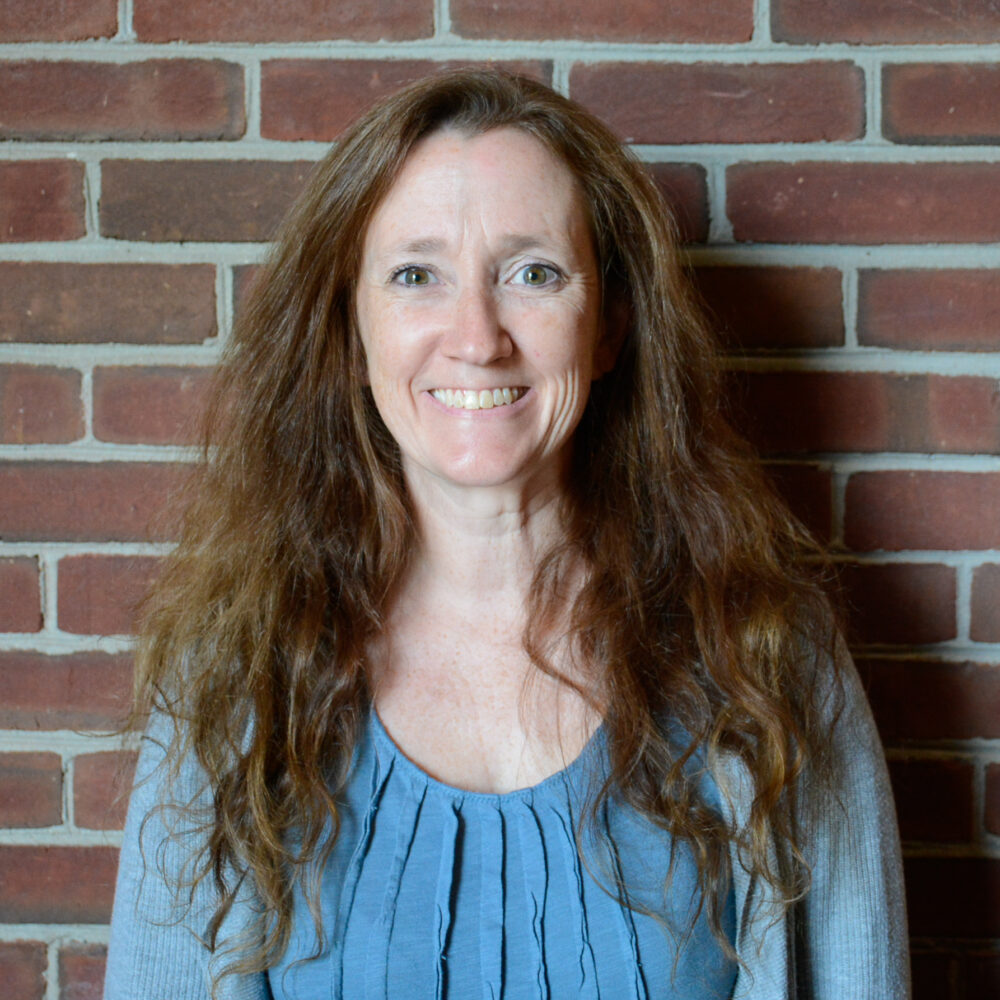 Smiling woman with long brown hair wearing a blue top, standing in front of a brick wall.