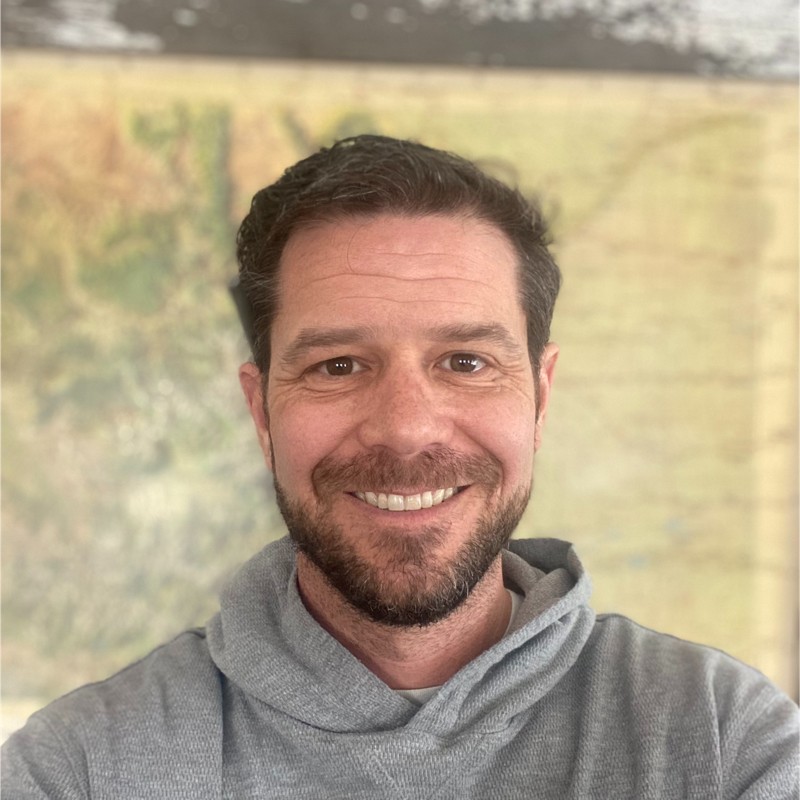 Portrait of a smiling man with short brown hair and a beard, wearing a gray hoodie, looking at the camera.