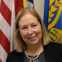 Portrait of a smiling woman with shoulder-length blond hair, wearing a dark blazer and pearl necklace, with the U.S. flag in the background.