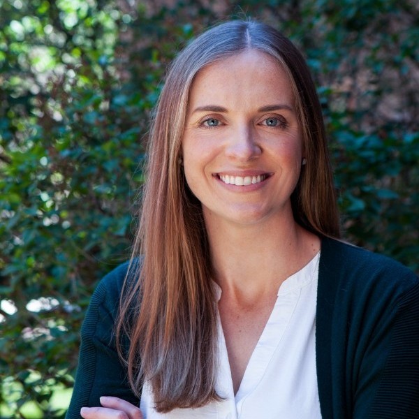 Smiling woman with long light brown hair in a white blouse and dark cardigan, outdoors with greenery in the background.