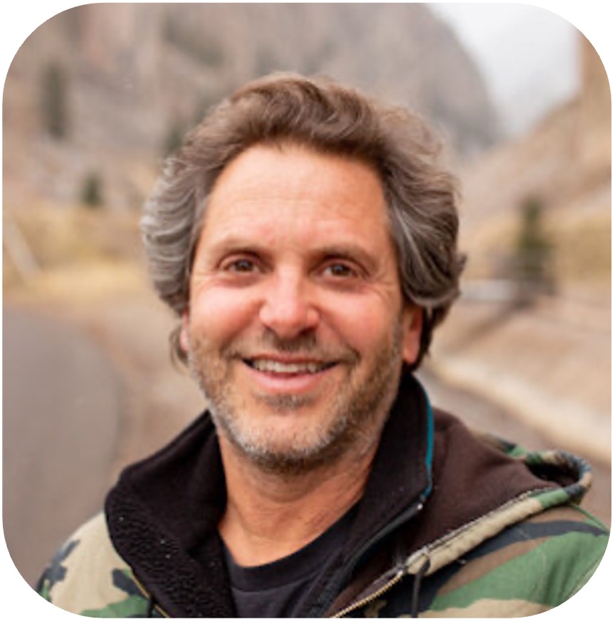 Smiling man with gray hair and stubble wearing a camouflage jacket, outdoors on a mountain road.