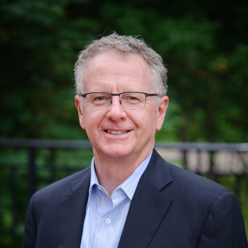 Portrait of a smiling man in a navy blazer and light blue striped shirt, wearing glasses, outdoors with a green, blurred background.