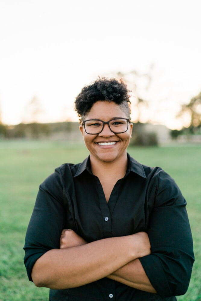 Smiling person with short curly hair and glasses, arms crossed, wearing a black button-down shirt outdoors in a grassy field.