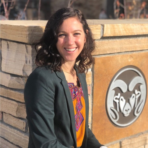 Smiling woman with dark hair in a blazer and orange embroidered top, seated beside a stone wall with a circular ram logo behind her.
