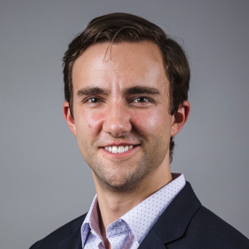 Head-and-shoulders portrait of a smiling man in a navy blazer and light polka-dot shirt against a gray background.