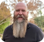 Smiling bald man with a long gray beard standing outdoors in a park with autumn foliage.