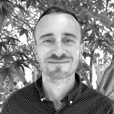 Portrait of a smiling man with short hair and a light beard, wearing a black polka-dot shirt, outdoors with leafy trees in the background.