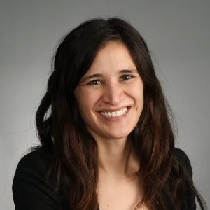 Portrait of a smiling woman with long dark hair wearing a black top, against a neutral gray background.