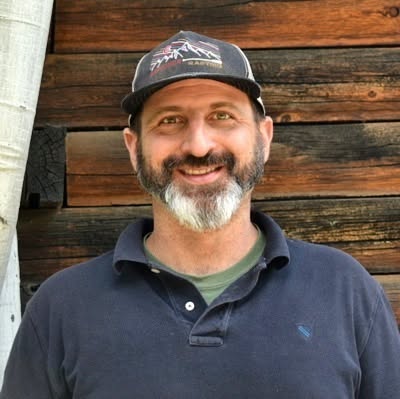 Smiling man with a gray beard in a navy polo shirt and cap, standing in front of a stacked wooden wall.