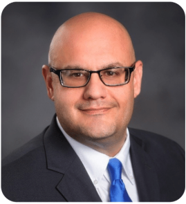Professional headshot of a bald man wearing glasses, a dark suit, white shirt, and blue tie, facing the camera with a neutral gray studio background.