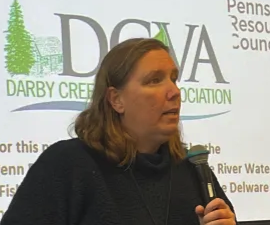 Woman with shoulder-length hair speaking into a microphone at a Darby Creek Association event, DCVA logo on the backdrop.