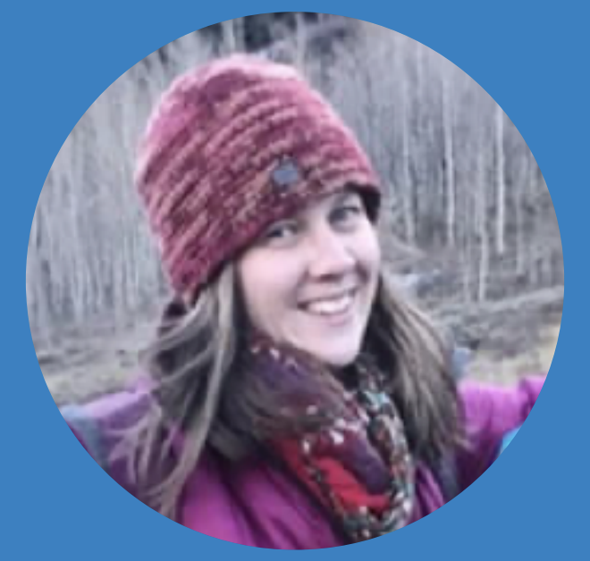Smiling woman outdoors wearing a red knit hat and purple jacket, with a scarf, in a bare-wooded background.