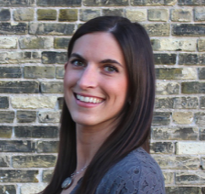 Smiling woman with long dark hair in a gray knit sweater standing against a brick wall.