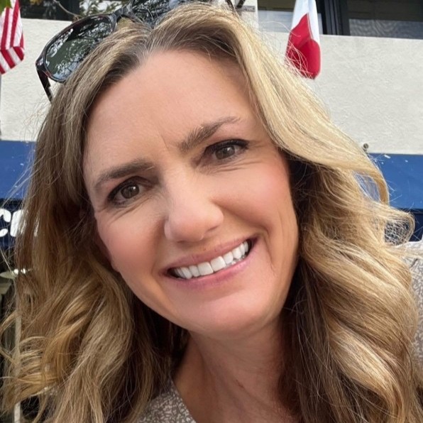 Close-up of a smiling woman with wavy blonde hair outdoors, sunglasses resting on her head.