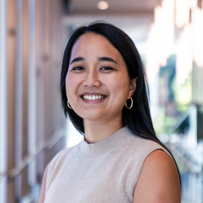 Portrait of a smiling woman with long dark hair wearing a beige knit sleeveless top and gold hoop earrings in a bright hallway