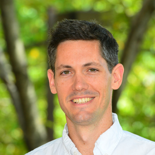 Smiling man with short dark hair in a white shirt outdoors, with green foliage in the background.