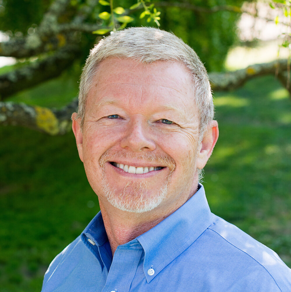 Smiling man with short gray hair and facial hair wearing a blue button-down shirt outdoors in a green setting.