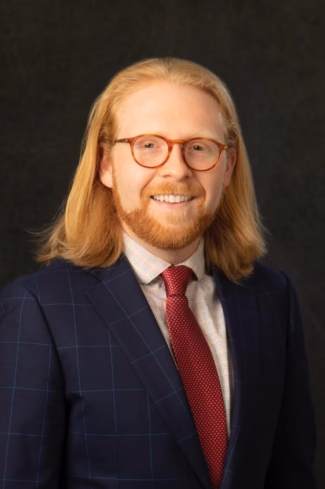 Portrait of a man with shoulder-length blond hair, red beard, and round glasses wearing a navy check suit and red tie against a dark background.