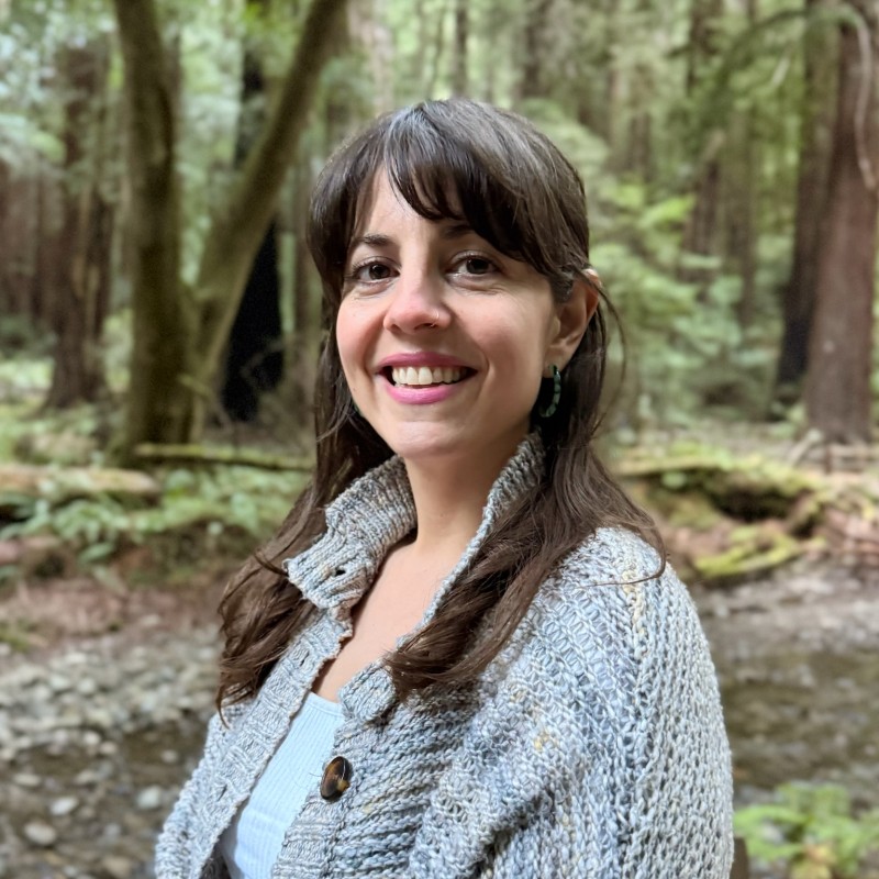 Smiling woman with brown hair in a gray knitted cardigan outdoors in a forest beside a creek.