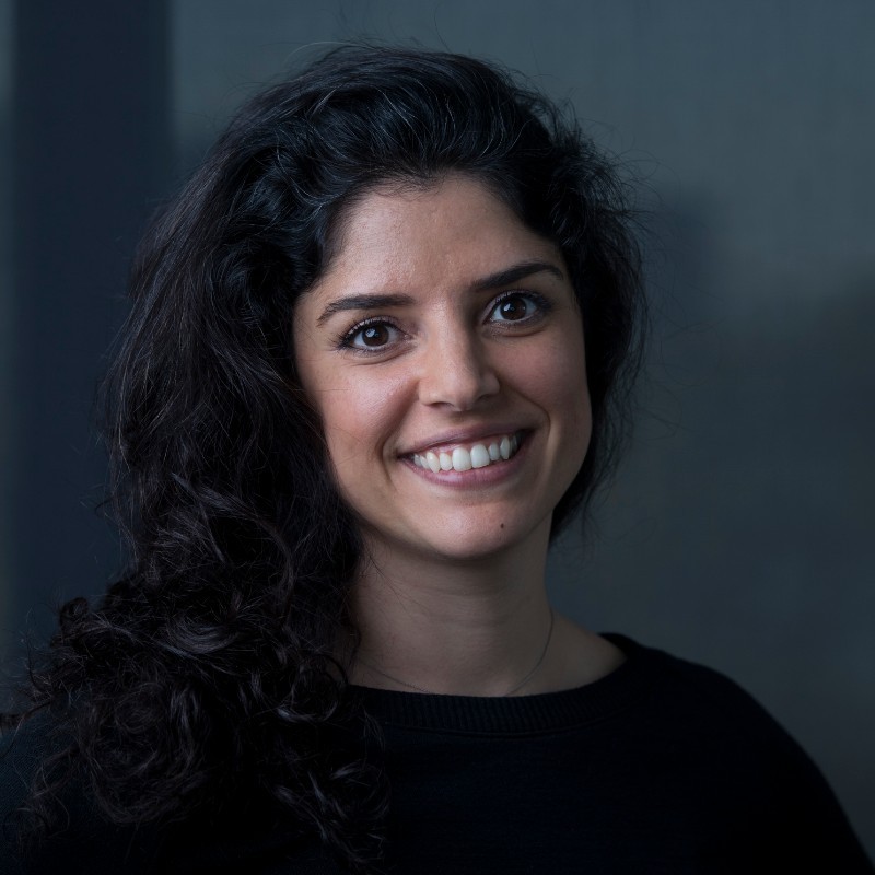 Smiling woman with dark curly hair and a black top against a dark gray background, looking at the camera.