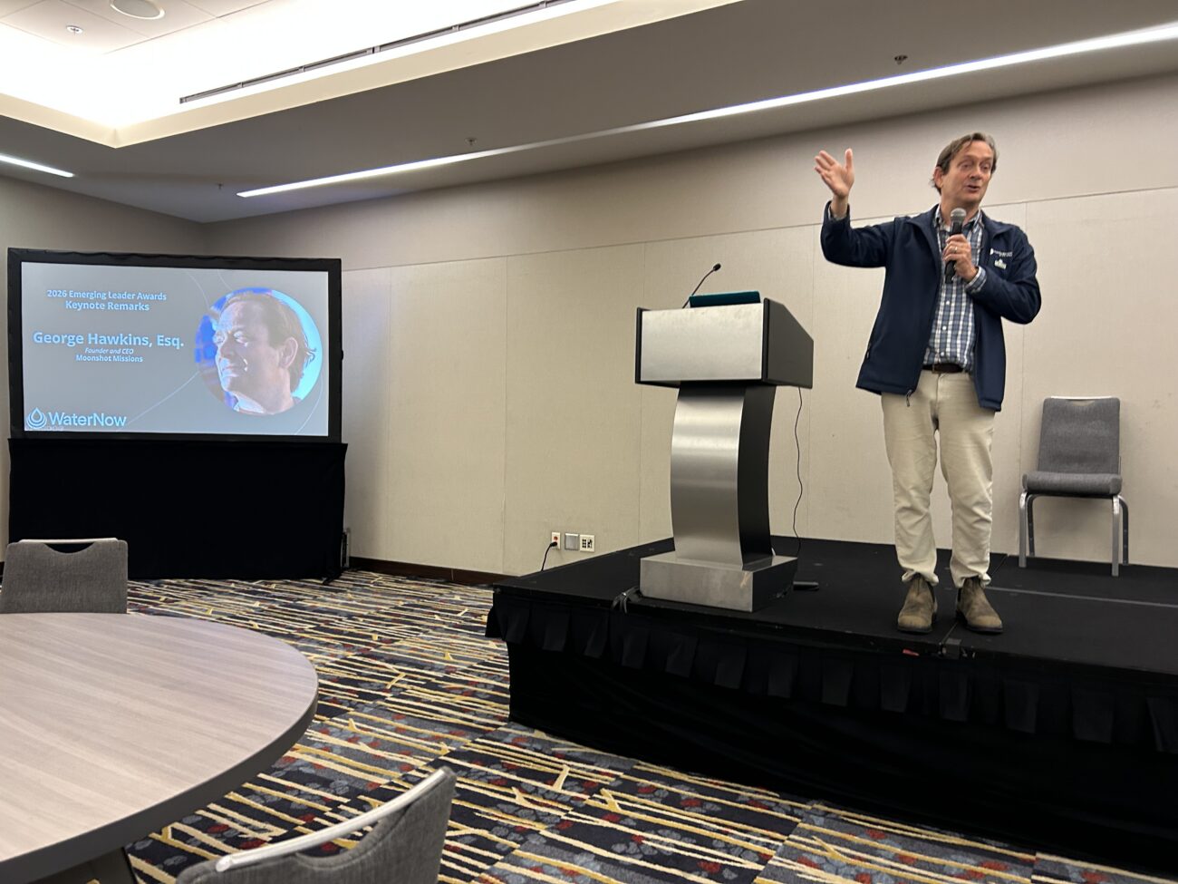 Speaker on stage holding a microphone and gesturing with one hand; podium and a projected slide are to the left showing a WaterNow keynote title and image of George Hawkins, Esq.